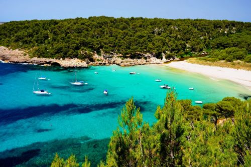The beach of Cala Trebaluger with its turquoise waters, lush vegetation and some boats at anchor